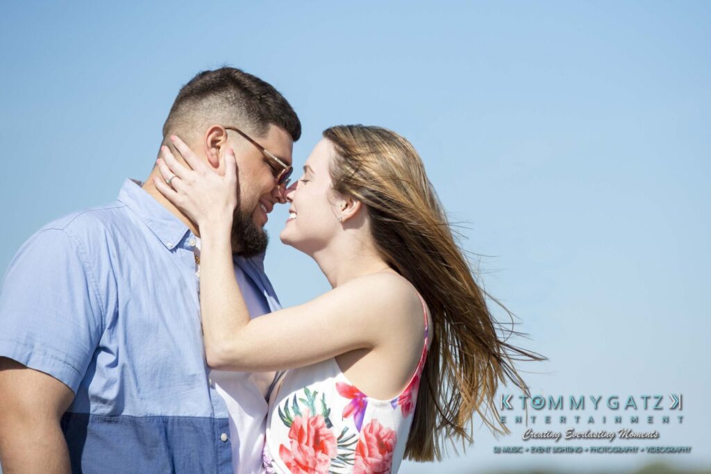 Kari-Jose-43018-FINALS-Low-Res-WM-23 - TOMMY GATZ ENTERTAINMENT A couple embraces and smiles against a clear blue sky. The woman wears a floral dress; the man wears a blue shirt.
