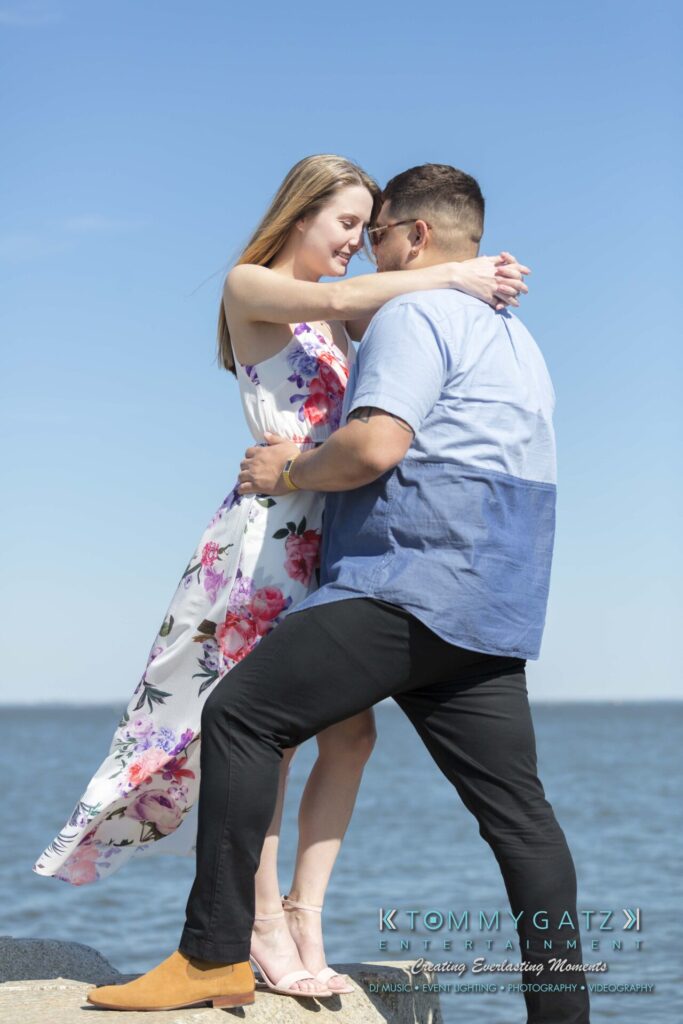 Kari-Jose-43018-FINALS-Low-Res-WM-27 - TOMMY GATZ ENTERTAINMENT A couple sharing a light moment on a rocky shore, with the ocean in the background. The woman wears a floral dress, and the man wears sunglasses and a two-tone shirt.