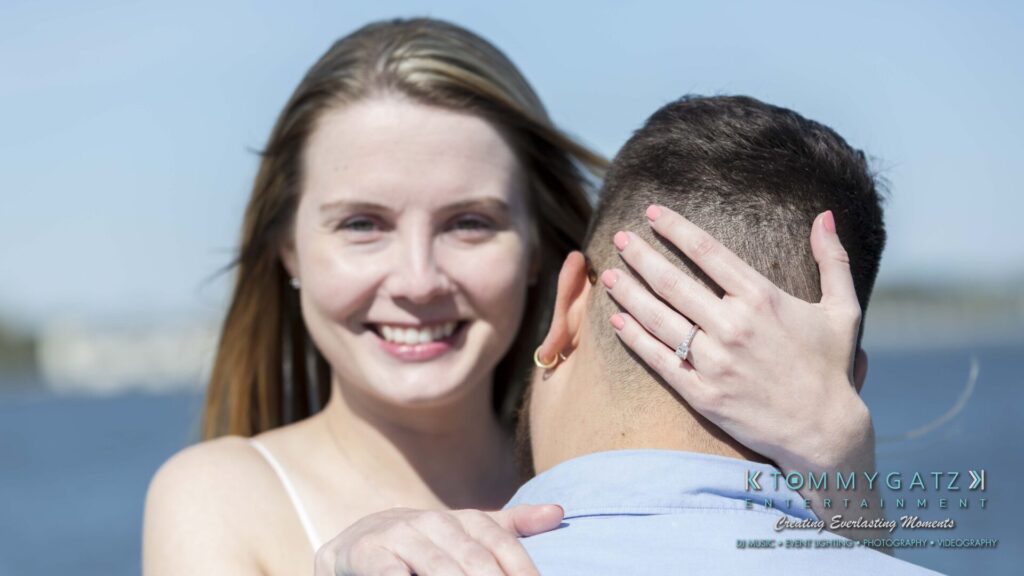 Kari-Jose-43018-FINALS-Low-Res-WM-29 - TOMMY GATZ ENTERTAINMENT A woman embraces a person while showing a ring on her hand. She is smiling, and they are outdoors near water, with the focus on her face.