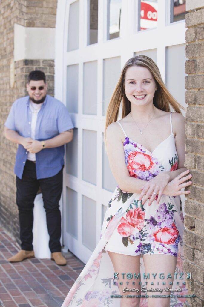 Kari-Jose-43018-FINALS-Low-Res-WM-32 - TOMMY GATZ ENTERTAINMENT A woman in a floral dress smiles at the camera while a man in a blue shirt stands in the background against a brick wall.