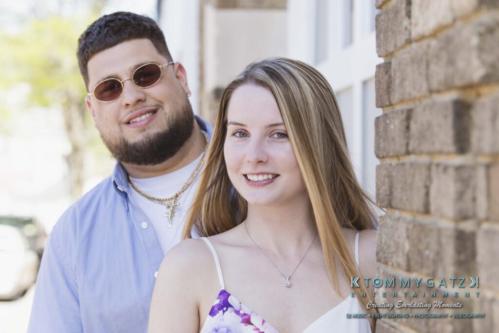 Kari-Jose-43018-FINALS-Low-Res-WM-33 - TOMMY GATZ ENTERTAINMENT A man and a woman stand closely together outdoors, both smiling. The man wears sunglasses and a blue shirt, and the woman is in a white floral dress.