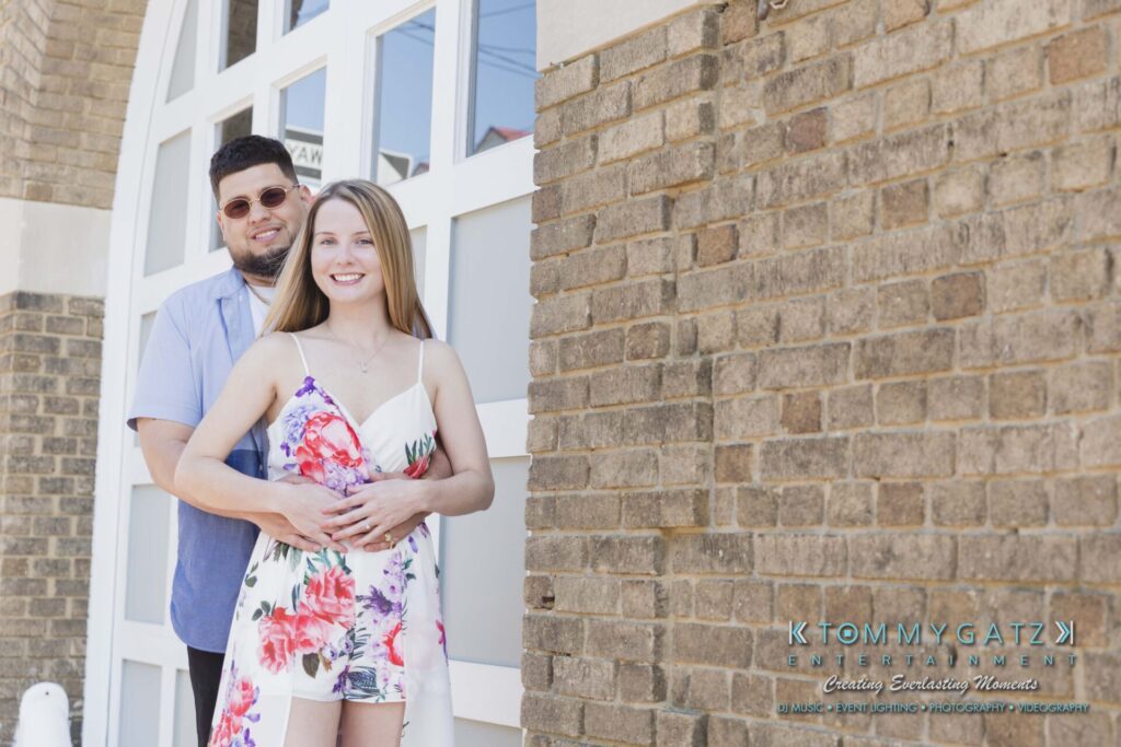 Kari-Jose-43018-FINALS-Low-Res-WM-35 - TOMMY GATZ ENTERTAINMENT A couple poses together in front of a brick building. The woman is wearing a floral dress, and the man is in a light shirt and sunglasses.