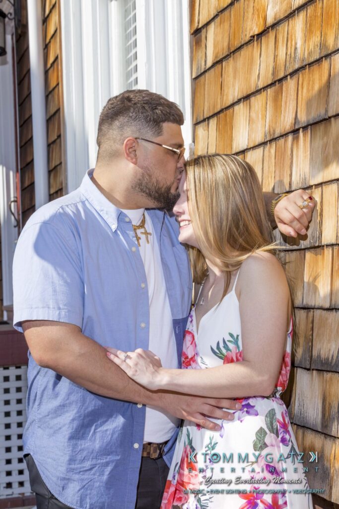 Kari-Jose-43018-FINALS-Low-Res-WM-38 - TOMMY GATZ ENTERTAINMENT A man in a blue shirt embraces a woman in a floral dress, standing close on a sunlit porch.