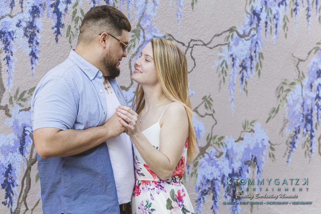 Kari-Jose-43018-FINALS-Low-Res-WM-42-1024x683 - TOMMY GATZ ENTERTAINMENT A couple holding hands and smiling at each other in front of a wall with painted blue flowers.