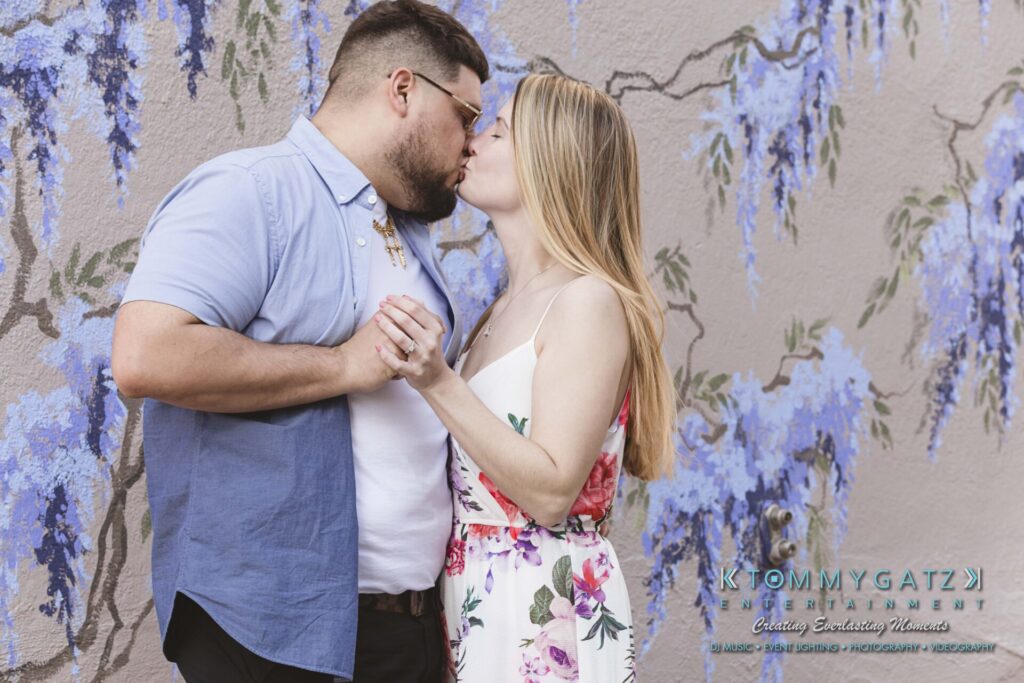 Kari-Jose-43018-FINALS-Low-Res-WM-43 - TOMMY GATZ ENTERTAINMENT A couple kisses while holding hands in front of a wall with purple wisteria flowers.