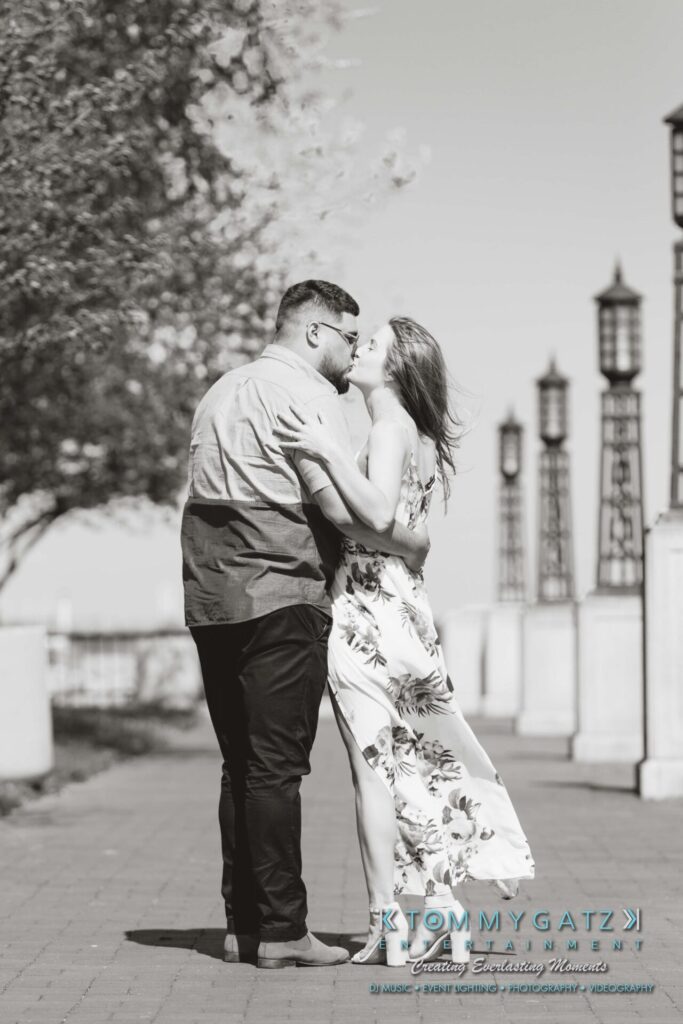 Kari-Jose-43018-FINALS-Low-Res-WM-6 - TOMMY GATZ ENTERTAINMENT A couple embraces while standing on a paved path lined with trees and lampposts. The woman wears a floral dress, and the man wears a short-sleeved shirt.