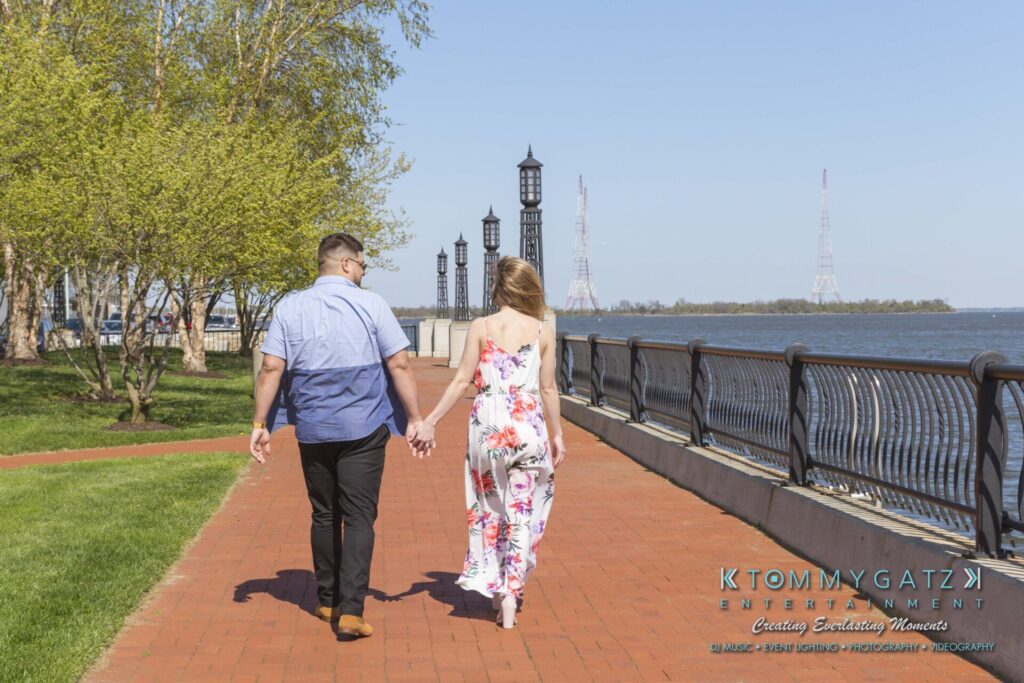Kari-Jose-43018-FINALS-Low-Res-WM-7 - TOMMY GATZ ENTERTAINMENT A couple holding hands walks along a brick path beside a waterfront with lamp posts and towers visible in the background.