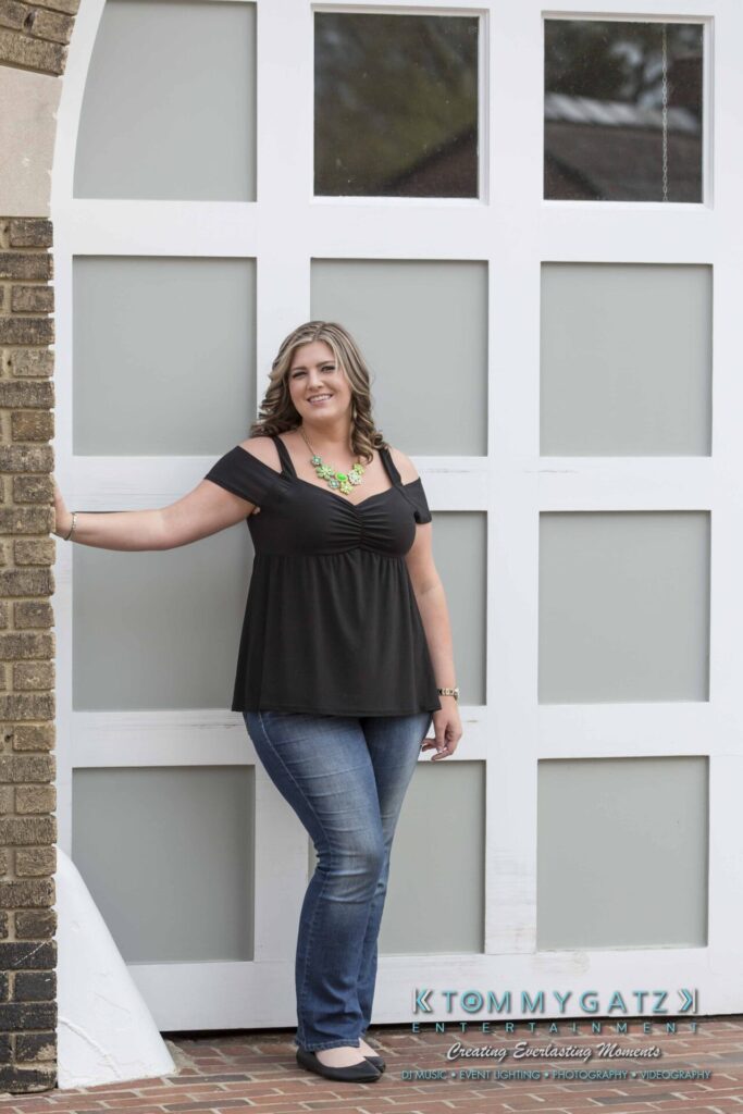 Lisa-Robert-Engagement-042218-Low-Res-Watermark-1 - TOMMY GATZ ENTERTAINMENT A woman in a black top and jeans stands against a large white door, smiling.
