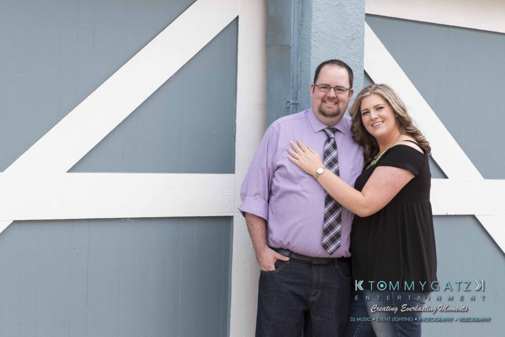 Lisa-Robert-Engagement-042218-Low-Res-Watermark-10 - TOMMY GATZ ENTERTAINMENT A man and woman pose together against a blue and white barn door. The woman rests her hand on the man's shoulder. Both are smiling.
