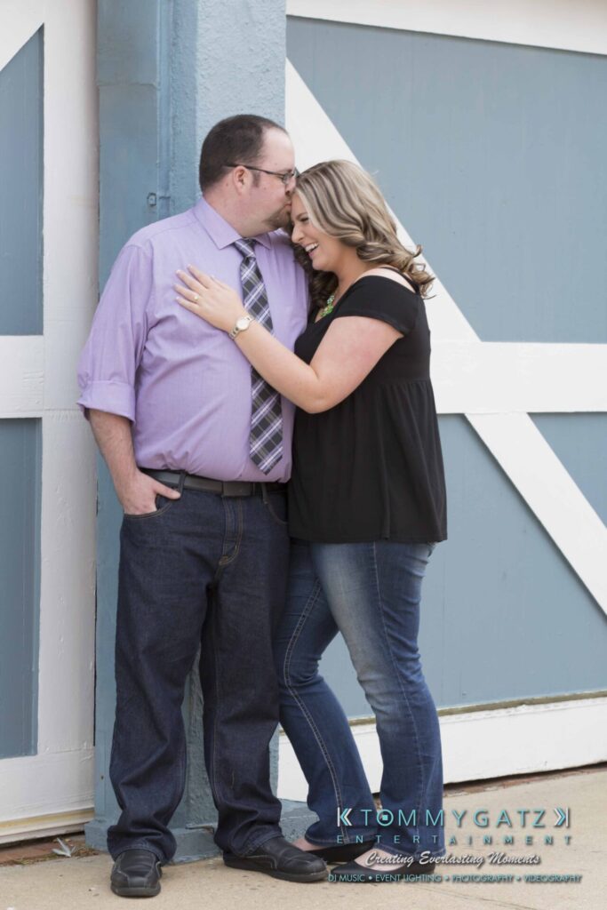 Lisa-Robert-Engagement-042218-Low-Res-Watermark-12 - TOMMY GATZ ENTERTAINMENT A couple stands outside, leaning against a blue and white door. The man wears a purple shirt and jeans, while the woman wears a black top and jeans. Both are smiling and embracing.