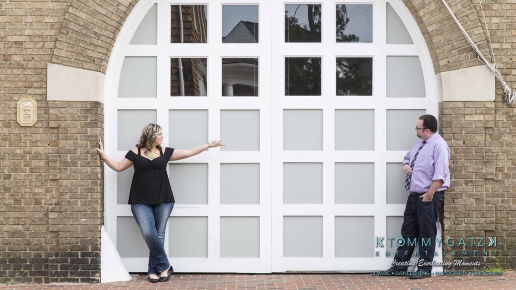 Lisa-Robert-Engagement-042218-Low-Res-Watermark-2 - TOMMY GATZ ENTERTAINMENT Two people stand apart, facing each other in front of a large arched window set in a brick wall. The person on the left points toward the right.