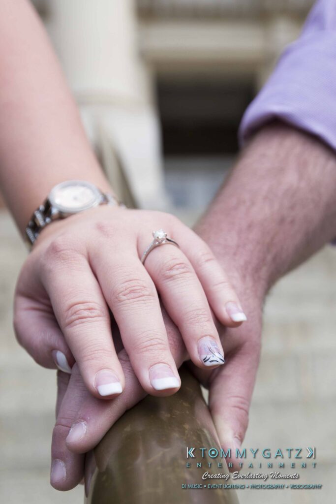 Lisa-Robert-Engagement-042218-Low-Res-Watermark-27 - TOMMY GATZ ENTERTAINMENT A close-up of two hands holding a metal railing. One hand has a watch and wears a ring with a large stone. The other hand rests beneath. Logo text is visible at the bottom.