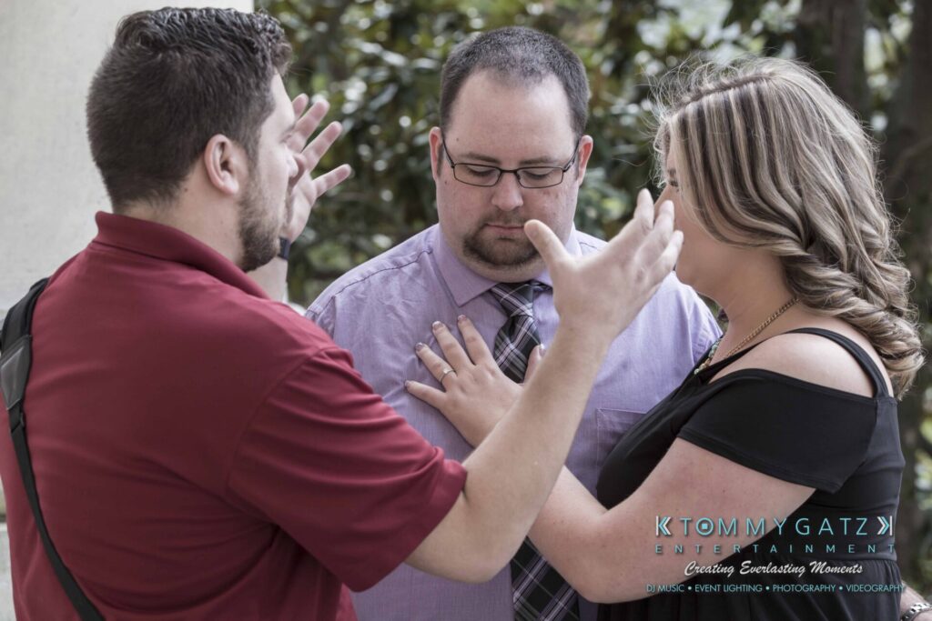 Lisa-Robert-Engagement-042218-Low-Res-Watermark-28 - TOMMY GATZ ENTERTAINMENT Three people are engaged in a conversation outdoors; a man in a red shirt gestures with his hands while a man in a purple shirt and a woman look on.