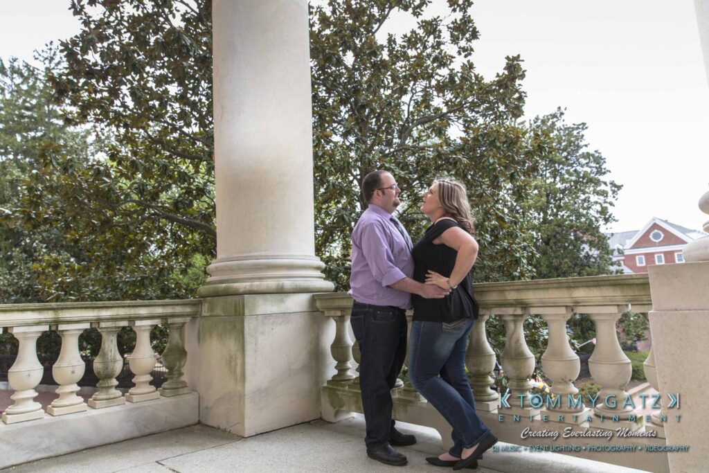 Lisa-Robert-Engagement-042218-Low-Res-Watermark-29 - TOMMY GATZ ENTERTAINMENT A couple stands on an outdoor balcony in front of a large tree, facing and embracing each other.