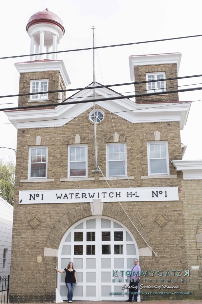 Lisa-Robert-Engagement-042218-Low-Res-Watermark-3 - TOMMY GATZ ENTERTAINMENT Old firehouse with a brick facade and arched garage. Two people stand in front. Sign reads "Waterwitch H-L No 1.