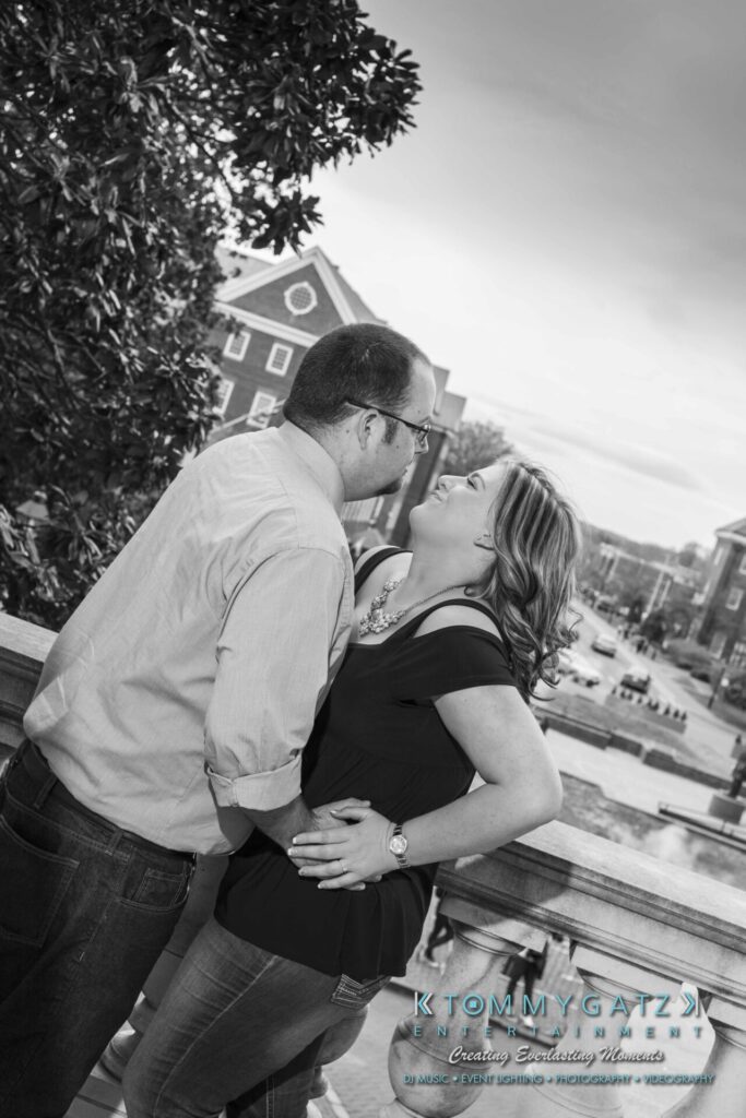 Lisa-Robert-Engagement-042218-Low-Res-Watermark-30 - TOMMY GATZ ENTERTAINMENT A couple stands on a balcony overlooking a street, embracing and smiling at each other.