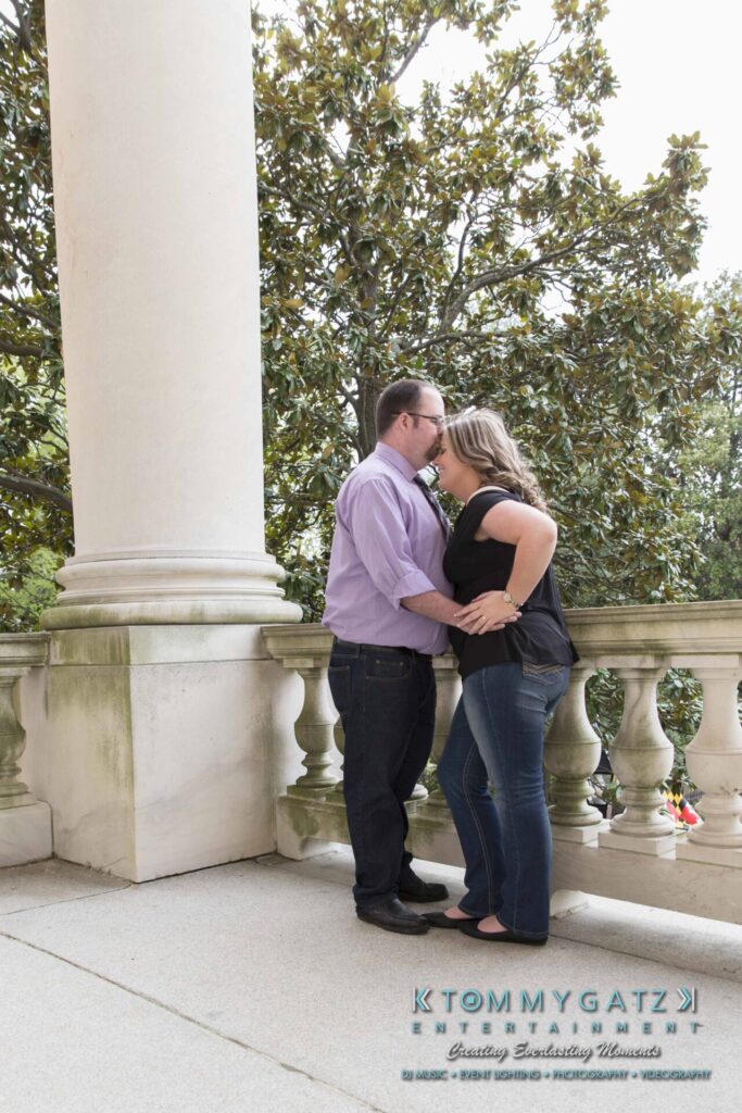 Lisa-Robert-Engagement-042218-Low-Res-Watermark-32 - TOMMY GATZ ENTERTAINMENT A couple stands on a balcony with white columns and greenery in the background, embracing closely.