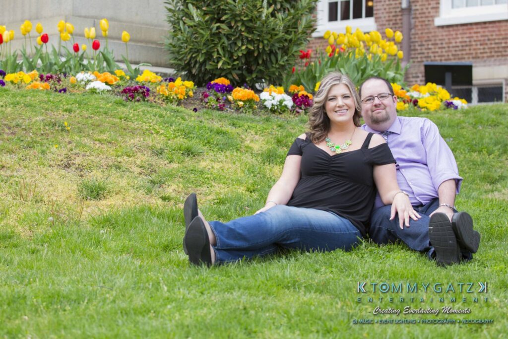 Lisa-Robert-Engagement-042218-Low-Res-Watermark-33 - TOMMY GATZ ENTERTAINMENT A couple sitting on grass in front of a building and colorful flower bed, smiling at the camera.