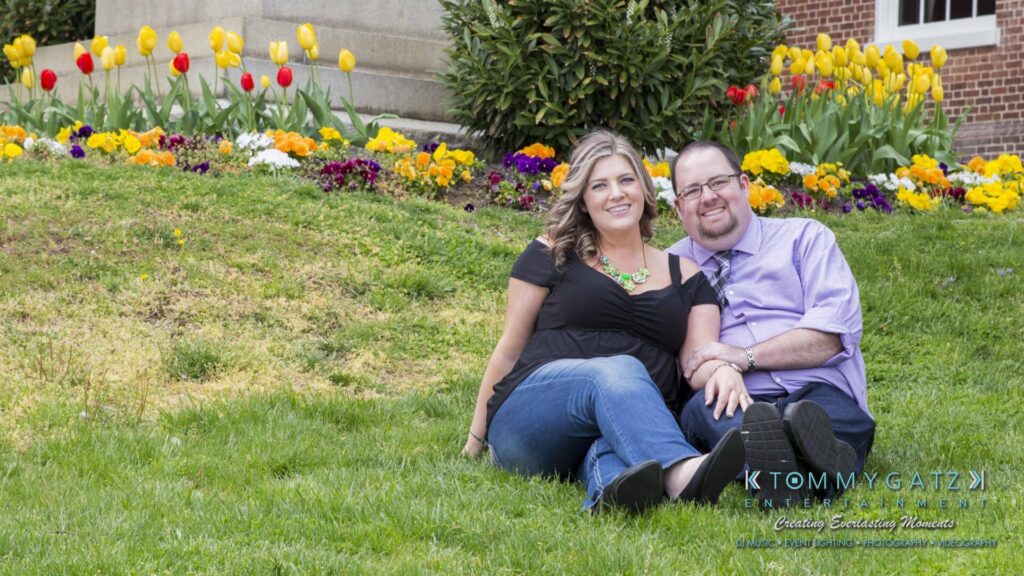 Lisa-Robert-Engagement-042218-Low-Res-Watermark-35 - TOMMY GATZ ENTERTAINMENT A couple sitting on a grassy lawn in front of colorful flower beds. The woman is wearing a black top and jeans, and the man is in a purple shirt and gray pants.