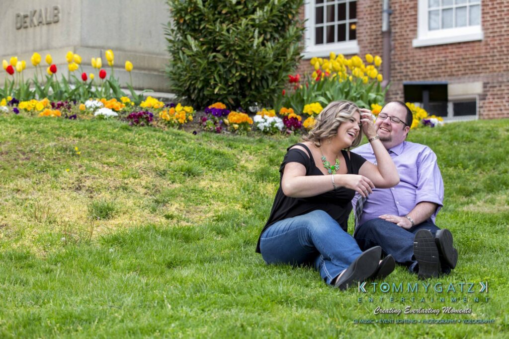 Lisa-Robert-Engagement-042218-Low-Res-Watermark-36 - TOMMY GATZ ENTERTAINMENT A woman and a man sit laughing on a grassy lawn with colorful flowers and a building in the background.