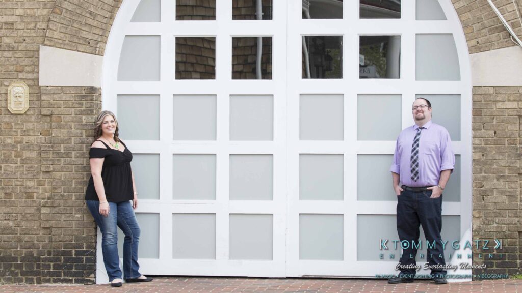 Lisa-Robert-Engagement-042218-Low-Res-Watermark-4 - TOMMY GATZ ENTERTAINMENT Two people standing in front of a large white door with frosted glass panels, posed on either side, both wearing casual attire.