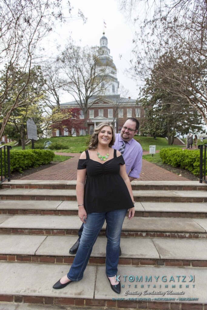 Lisa-Robert-Engagement-042218-Low-Res-Watermark-41 - TOMMY GATZ ENTERTAINMENT A smiling woman and man pose on stone steps, with a historic building and trees in the background.