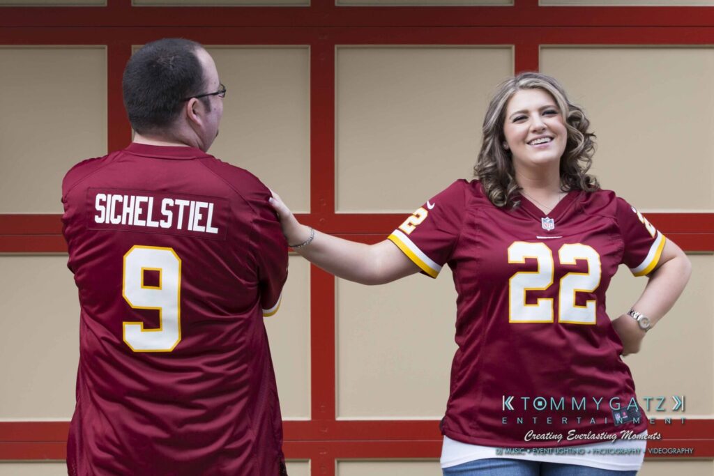 Lisa-Robert-Engagement-042218-Low-Res-Watermark-46 - TOMMY GATZ ENTERTAINMENT A woman in a red sports jersey, number 22, smiles while extending her hand to a man in a red jersey, number 9, with "Sichelstiel" written on it. They stand in front of a red grid-patterned wall.