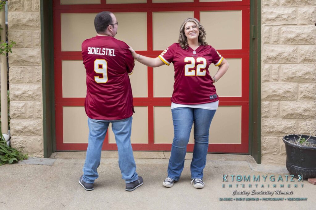 Lisa-Robert-Engagement-042218-Low-Res-Watermark-47 - TOMMY GATZ ENTERTAINMENT Two people wearing matching red sports jerseys stand outside a garage door. The woman holds the man's hand while both smile.