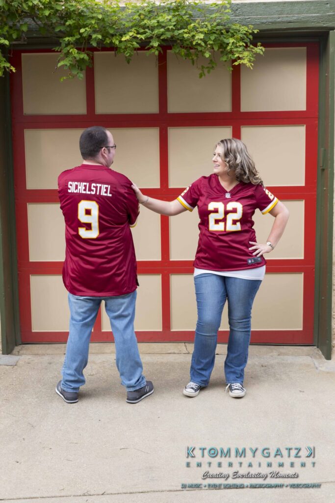 Lisa-Robert-Engagement-042218-Low-Res-Watermark-48 - TOMMY GATZ ENTERTAINMENT Two people in matching red sports jerseys stand in front of a garage. The man shows the number 9, and the woman shows the number 22 on their jerseys.