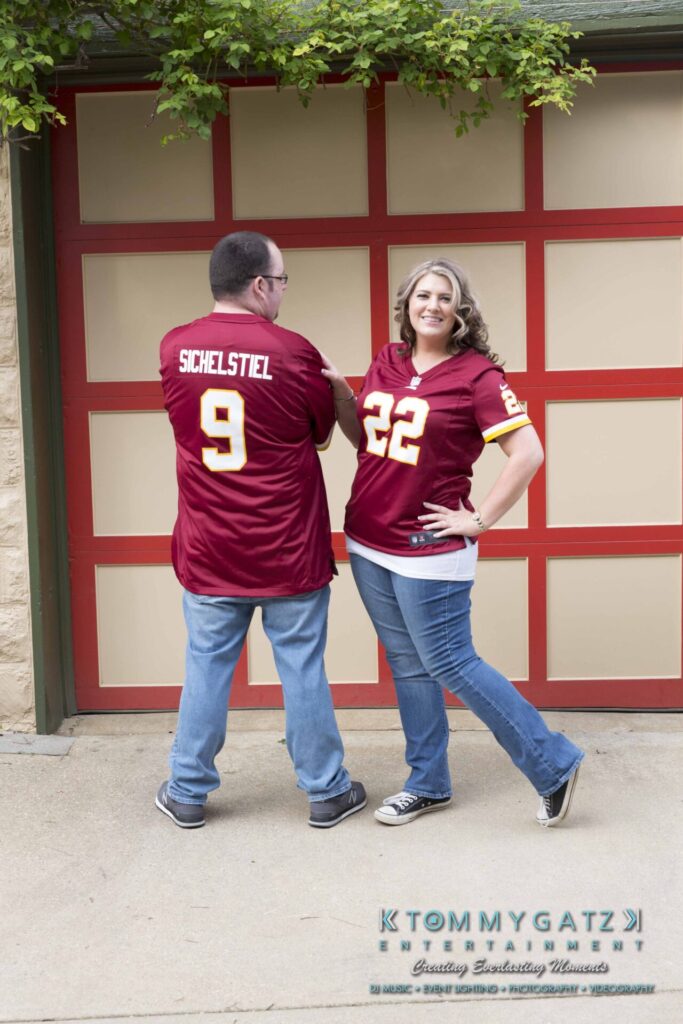 Lisa-Robert-Engagement-042218-Low-Res-Watermark-49 - TOMMY GATZ ENTERTAINMENT Two people wearing matching sports jerseys, numbered 9 and 22, stand in front of a red and white garage door. The man points to the name on his jersey, while the woman smiles with hands on hips.
