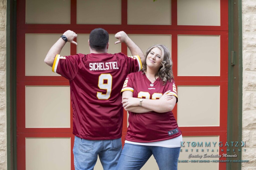 Lisa-Robert-Engagement-042218-Low-Res-Watermark-50 - TOMMY GATZ ENTERTAINMENT Two people wearing matching red sports jerseys stand in front of a brown garage door. The man shows the name "Sichelstiel" on his back, while the woman stands with crossed arms, smiling.