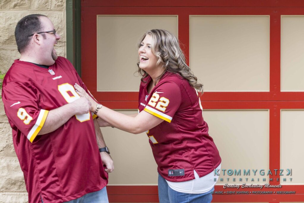 Lisa-Robert-Engagement-042218-Low-Res-Watermark-55 - TOMMY GATZ ENTERTAINMENT Two people wearing matching red sports jerseys are standing in front of a red and beige garage door, smiling and playfully interacting.