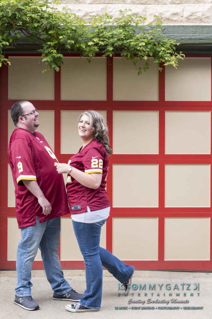 Lisa-Robert-Engagement-042218-Low-Res-Watermark-56 - TOMMY GATZ ENTERTAINMENT A man and woman wearing matching maroon sports jerseys stand in front of a red and beige garage door, smiling at each other.