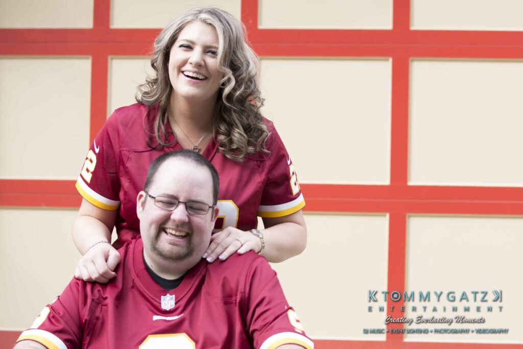 Lisa-Robert-Engagement-042218-Low-Res-Watermark-58 - TOMMY GATZ ENTERTAINMENT A woman and a man, both wearing red sports jerseys, are smiling. She stands behind him with hands on his shoulders.