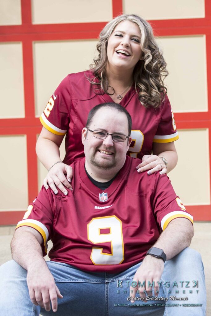 Lisa-Robert-Engagement-042218-Low-Res-Watermark-59 - TOMMY GATZ ENTERTAINMENT A woman and man wearing matching football jerseys pose together, with the woman standing behind the sitting man.
