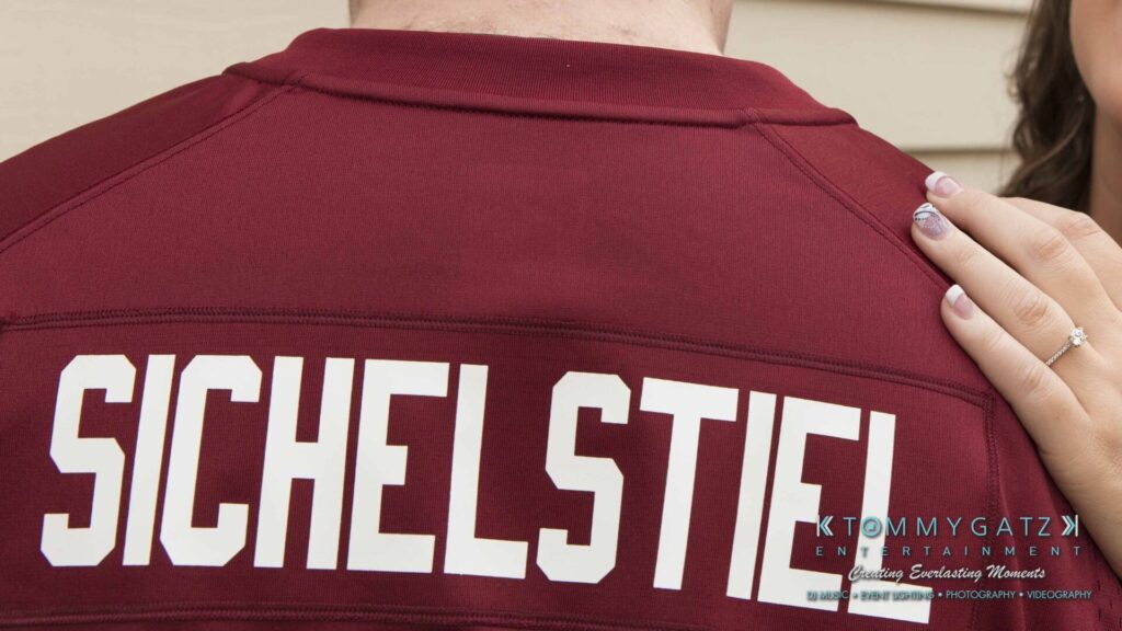 Lisa-Robert-Engagement-042218-Low-Res-Watermark-61 - TOMMY GATZ ENTERTAINMENT Back of a person wearing a maroon shirt with "Sichelstiel" printed on it, next to a hand resting on their shoulder.