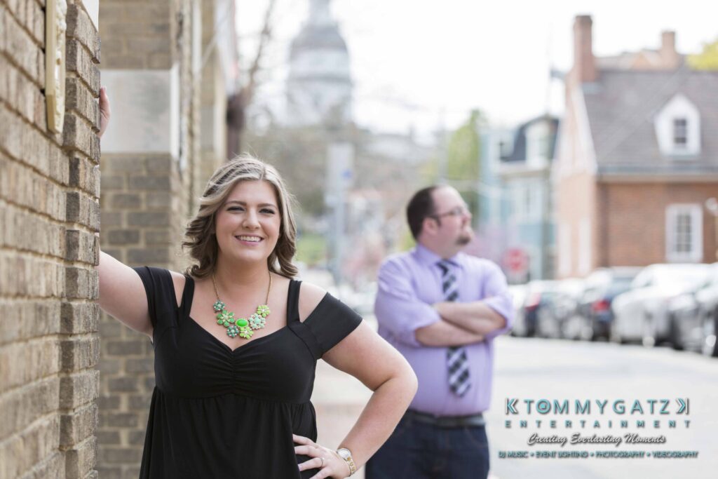 Lisa-Robert-Engagement-042218-Low-Res-Watermark-7 - TOMMY GATZ ENTERTAINMENT Woman in black dress leans against a brick wall, smiling, while a man in a purple shirt stands in the background with his arms crossed.