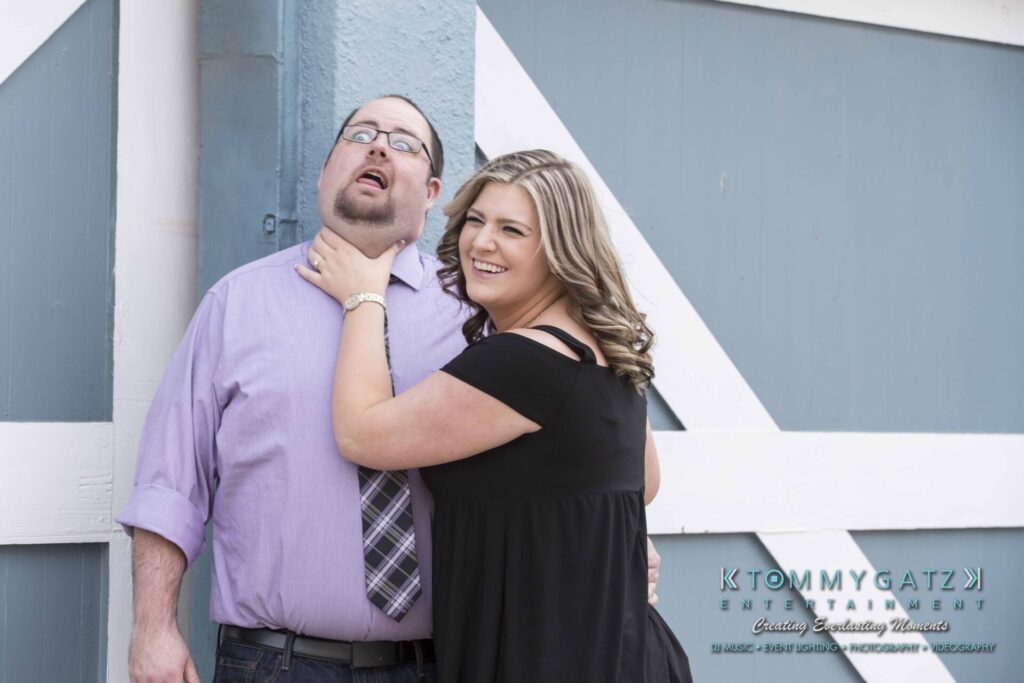 Lisa-Robert-Engagement-042218-Low-Res-Watermark-9 - TOMMY GATZ ENTERTAINMENT A woman playfully pretends to choke a man while both smile in front of a blue and white wall.