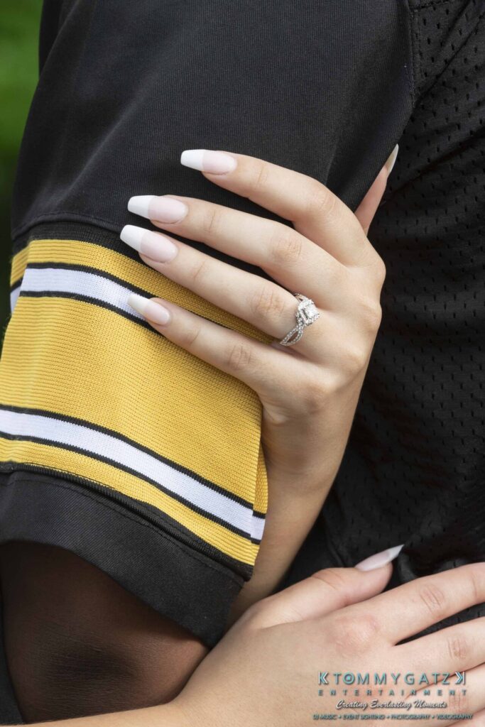 051919-Viona-Kevin-FINALS-Low-RES-WM-11 - TOMMY GATZ ENTERTAINMENT A hand with manicured nails rests on a person's arm, wearing a gold and black jersey. A diamond engagement ring is visible.