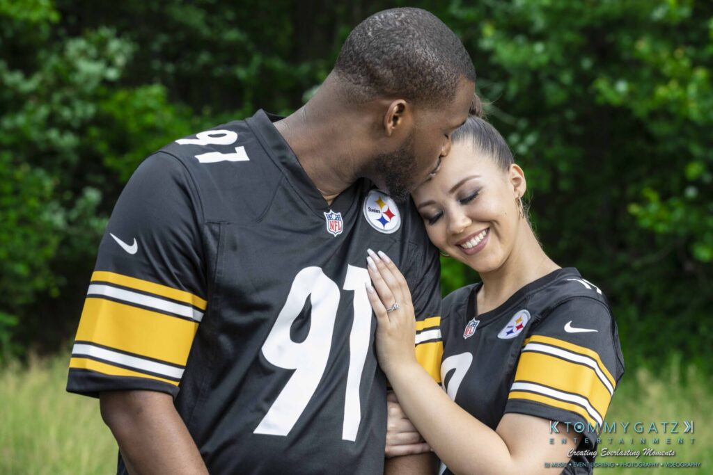 051919-Viona-Kevin-FINALS-Low-RES-WM-17 - TOMMY GATZ ENTERTAINMENT A couple wearing matching football jerseys, with the man kissing the woman's forehead. They are outdoors with greenery in the background.