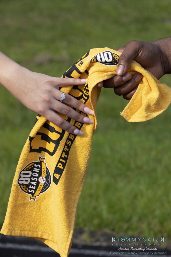 051919-Viona-Kevin-FINALS-Low-RES-WM-2 - TOMMY GATZ ENTERTAINMENT Two hands hold a yellow towel with a Pittsburgh Steelers design against a grassy background.