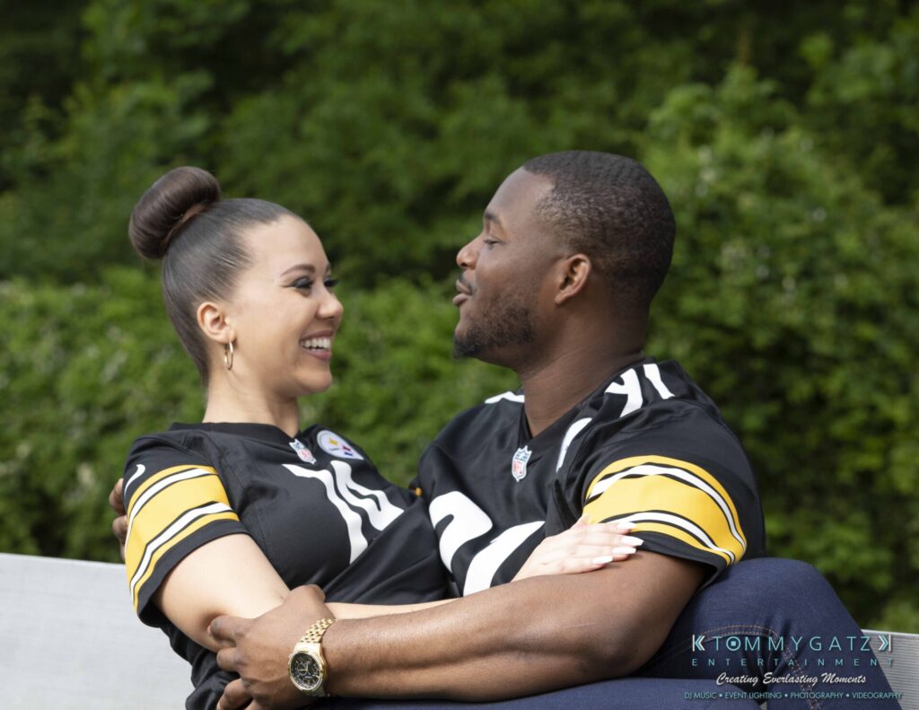 051919-Viona-Kevin-FINALS-Low-RES-WM-20 - TOMMY GATZ ENTERTAINMENT A couple wearing matching sports jerseys sits closely on a bench outdoors, smiling at each other against a lush green background.