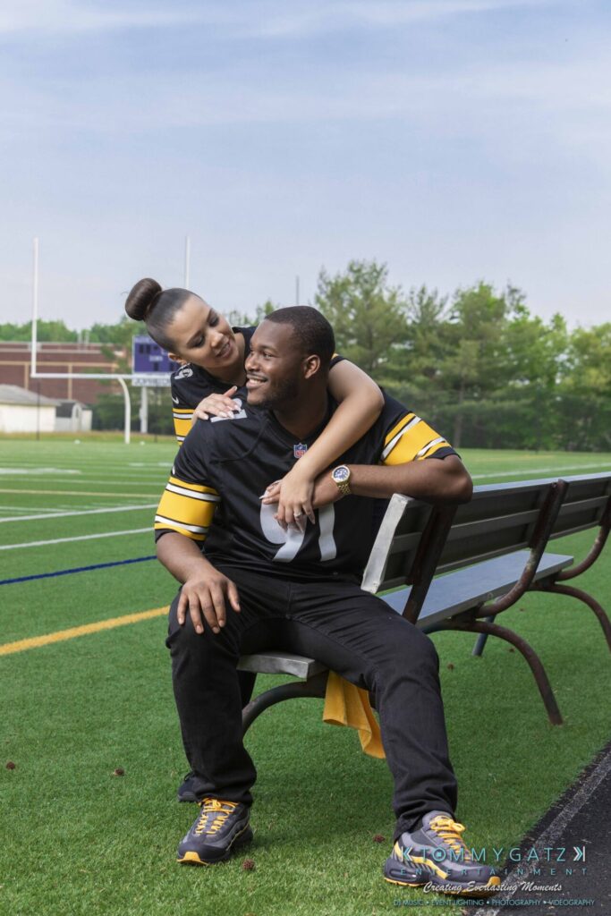 051919-Viona-Kevin-FINALS-Low-RES-WM-22 - TOMMY GATZ ENTERTAINMENT A couple wearing matching sports jerseys sit on a bench at a football field. The woman leans on the man's shoulders, smiling.
