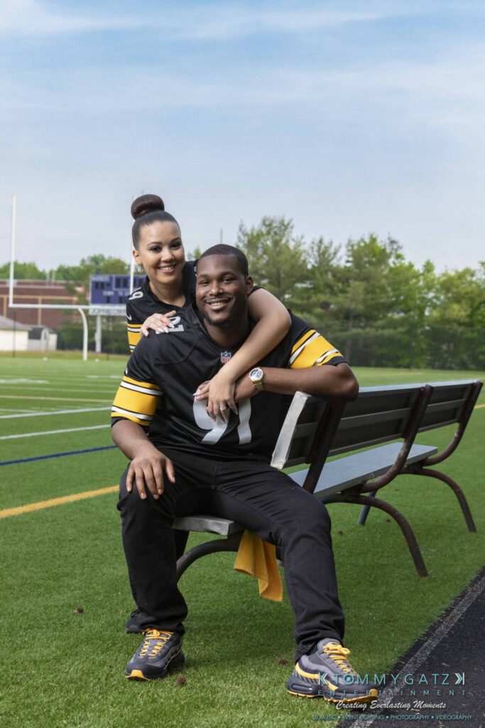 051919-Viona-Kevin-FINALS-Low-RES-WM-23 - TOMMY GATZ ENTERTAINMENT A couple wearing matching sports jerseys sits on a bench at a football field. The woman is smiling and has her arm around the man, who is also smiling.