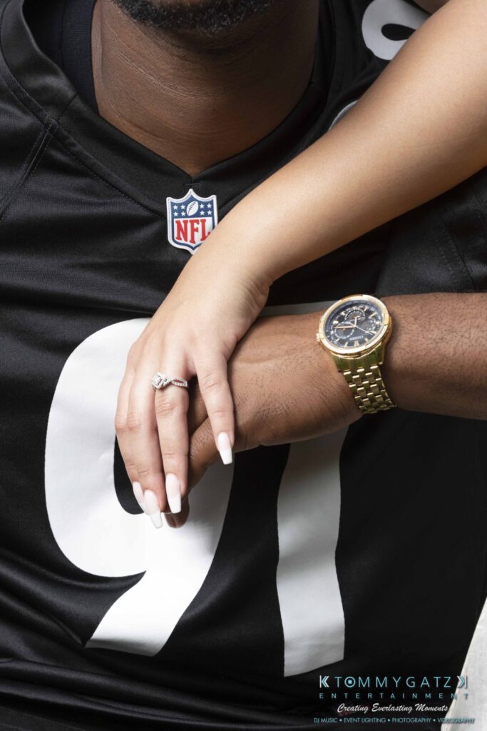 051919-Viona-Kevin-FINALS-Low-RES-WM-24 - TOMMY GATZ ENTERTAINMENT A person wearing a black NFL jersey and a gold watch with another person's hand resting on theirs, showcasing an engagement ring.