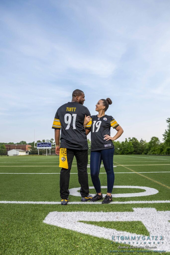 051919-Viona-Kevin-FINALS-Low-RES-WM-26 - TOMMY GATZ ENTERTAINMENT A couple stands on a football field wearing matching jerseys with numbers 91 and 19. The man holds a yellow towel.