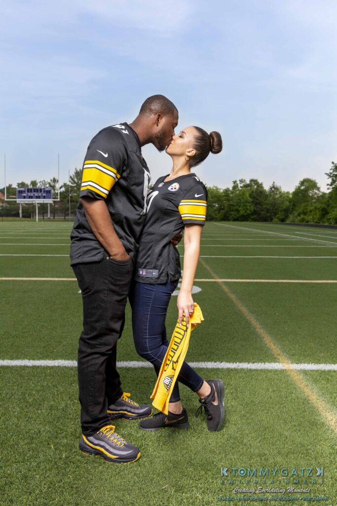 051919-Viona-Kevin-FINALS-Low-RES-WM-29 - TOMMY GATZ ENTERTAINMENT A couple in matching black and yellow sports jerseys kisses on a football field, with the woman holding a yellow towel.