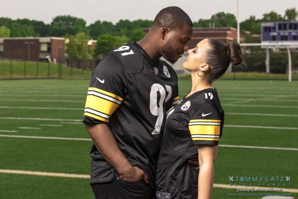 051919-Viona-Kevin-FINALS-Low-RES-WM-34 - TOMMY GATZ ENTERTAINMENT A couple in matching black football jerseys shares a moment on a field, with a school building in the background.