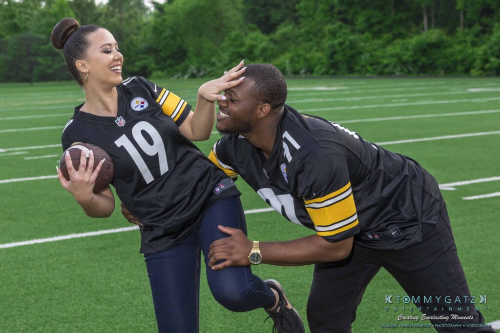 051919-Viona-Kevin-FINALS-Low-RES-WM-44 - TOMMY GATZ ENTERTAINMENT A woman holding a football playfully evades a man on a grassy field. Both wear black sports jerseys with yellow accents, numbered 19 and 91. Trees and a small structure are visible in the background.