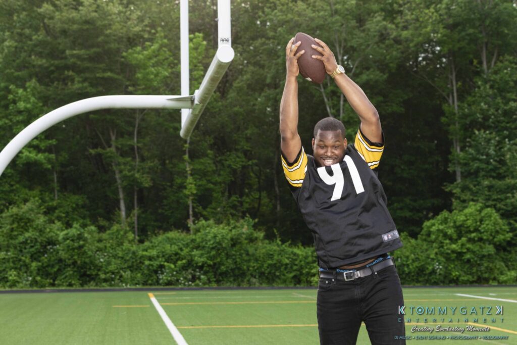 051919-Viona-Kevin-FINALS-Low-RES-WM-46 - TOMMY GATZ ENTERTAINMENT A man wearing a black and yellow sports jersey holds a football above his head on a football field near a goalpost.